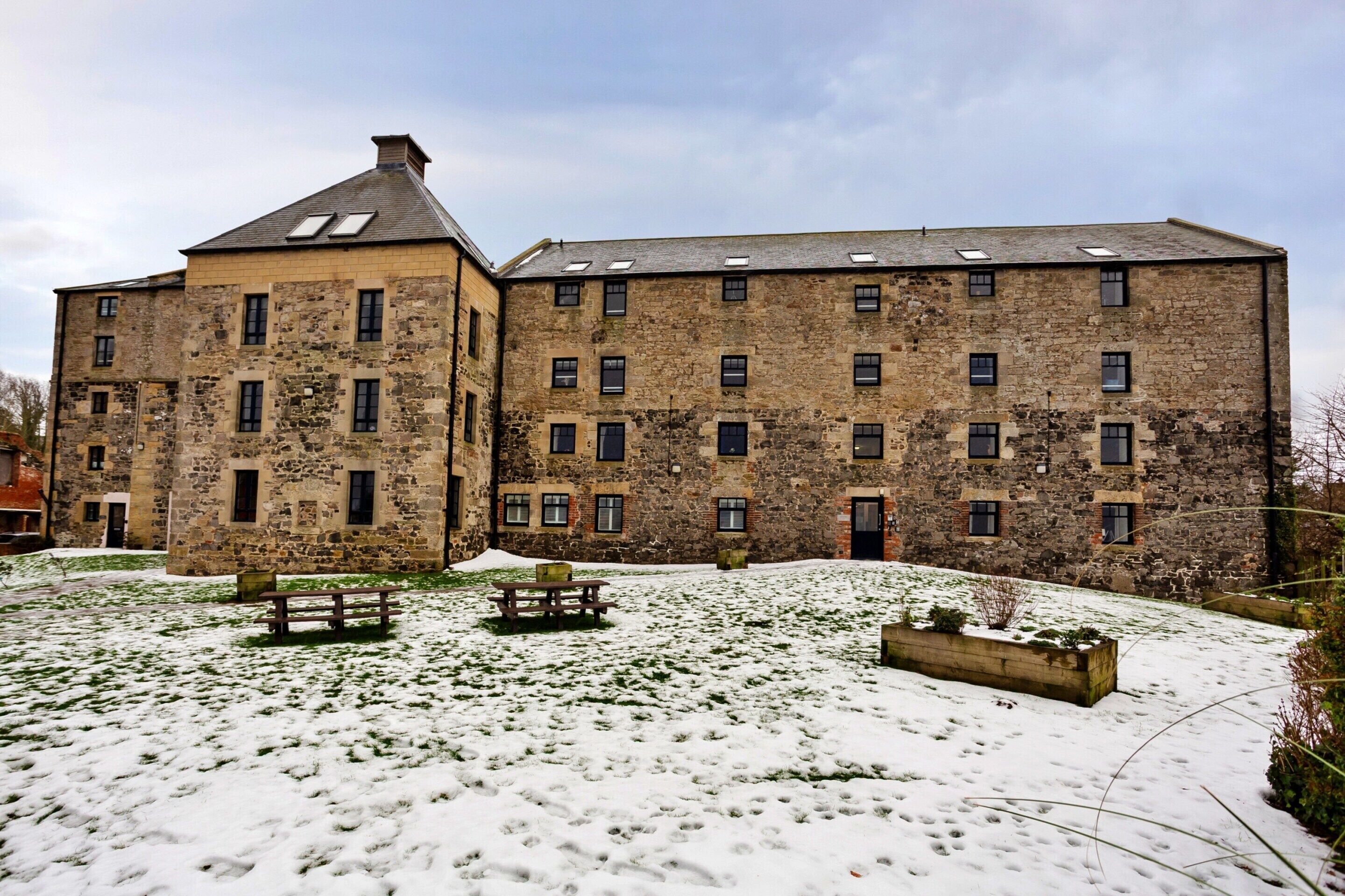 A stone building with multiple windows and a snow-covered courtyard with benches.