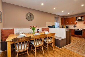 Un comedor con una mesa de madera dispuesta para cuatro, un reloj en la pared y una cocina al fondo.