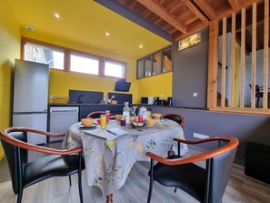 A dining area with a table set for two, a kitchen in the background, and a window with blinds. - Welcoming gîte, pets allowed - 5 guests (Doizieux)