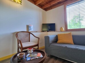 A room with a wooden ceiling, a grey sofa, a wicker chair, a small table with a mug and a brochure, a television on a stand, and a window with blinds. - Welcoming gîte, pets allowed - 5 guests (Doizieux)