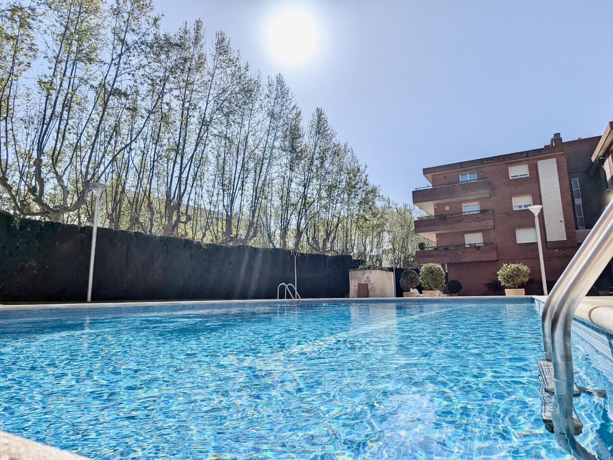 En swimmingpool med klart, blått vatten, omgiven av träd och en byggnad i bakgrunden.