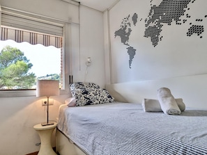 A bedroom with a bed, a lamp, a world map wall art, and a window with a view of trees. - Apartment with pool, pets OK - 6 guests (Tossa de Mar)