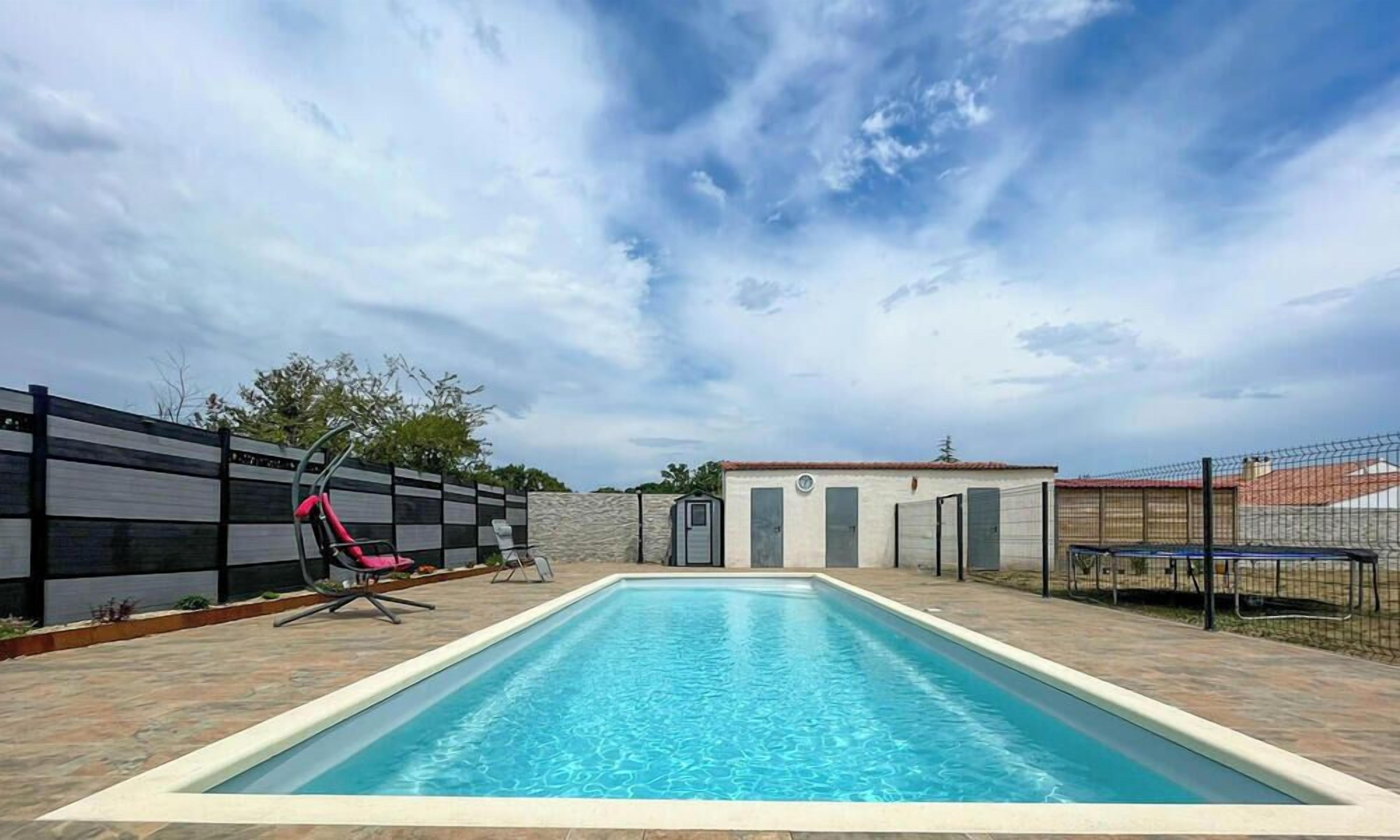 Una piscina rectangular con un borde de baldosas, una silla y un pequeño edificio con puerta y ventana.