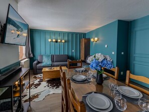 Ein Essbereich mit einem Holztisch, gedeckt für vier Personen, einer Couch und einem an der Wand befestigten Fernseher.