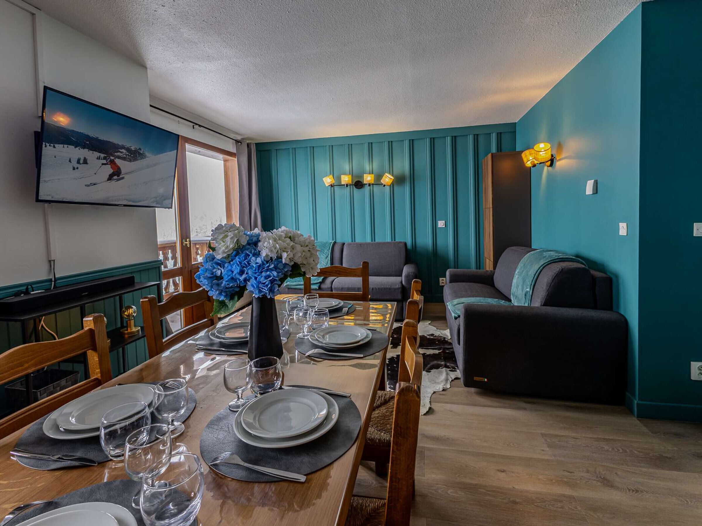 A dining area with a wooden table set for four, a vase of flowers, and a television mounted on the wall.