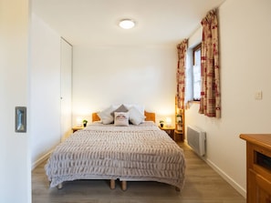Quarto individual com cama de solteiro, cabeceira de madeira, mesa de cabeceira com abajur, janela com cortinas e radiador.