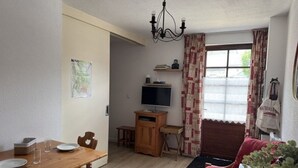 Uma área de refeições com uma mesa e cadeiras de madeira, uma televisão em um suporte, um sofá e uma janela com cortinas.