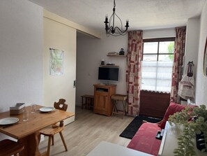 A dining area with a wooden table and chairs, a television on a stand, a sofa, and a window with curtains. - Cozy apartment - 4 guests (Vignec)