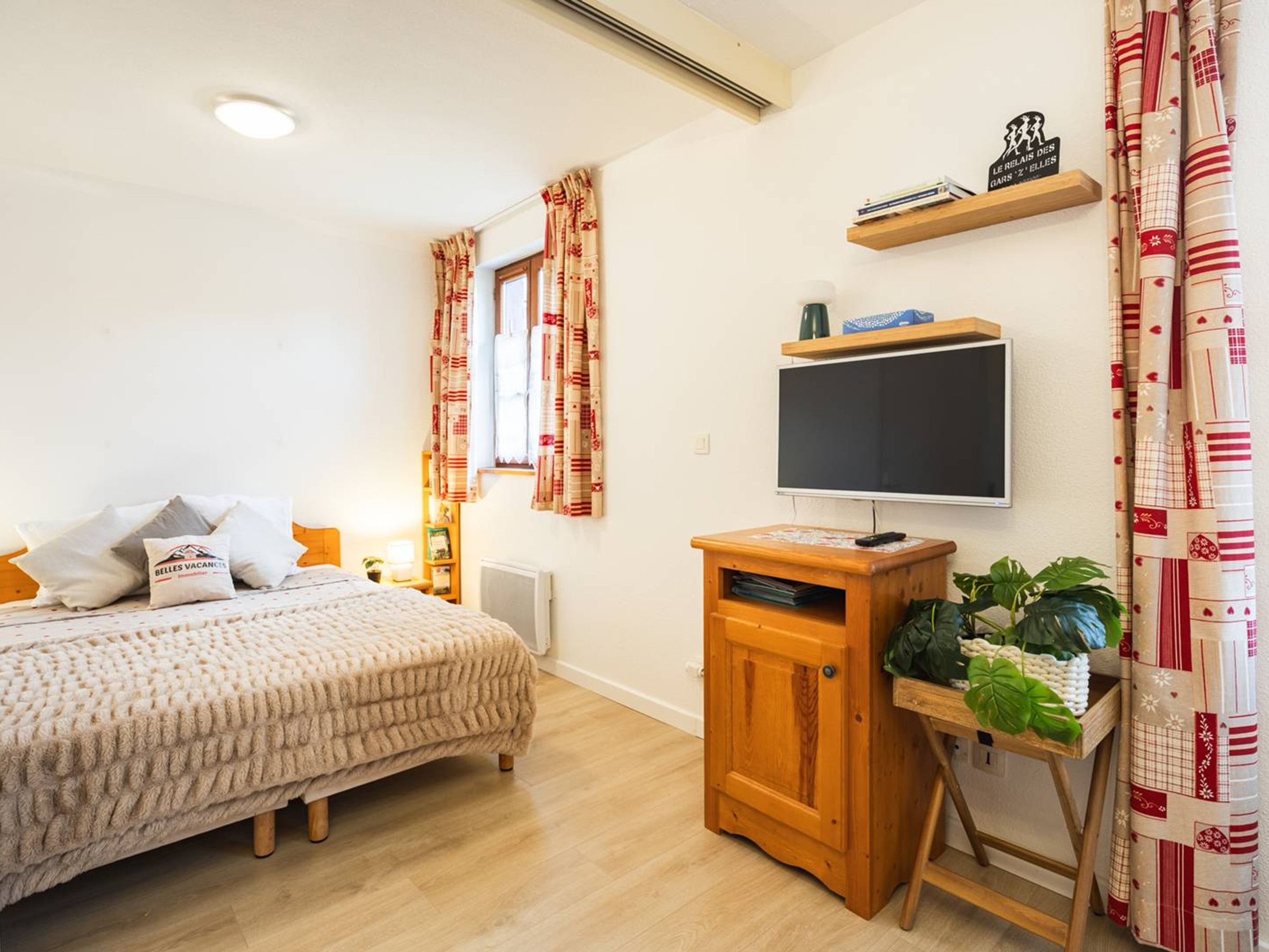 A bedroom with a bed, a TV on a wooden stand, a shelf with books, and a window with curtains.