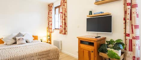 A bedroom with a bed, a TV on a wooden stand, a shelf with books, and a window with curtains.