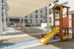 Ein Spielplatz mit Rutsche und Klettergerüst, umgeben von einem überdachten Bereich mit gemustertem Boden.