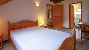 A bedroom with a wooden bed, a nightstand, a desk with a chair, and a door leading to another room.