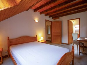 A bedroom with a wooden bed, a nightstand, a desk with a chair, and a door leading to another room.
