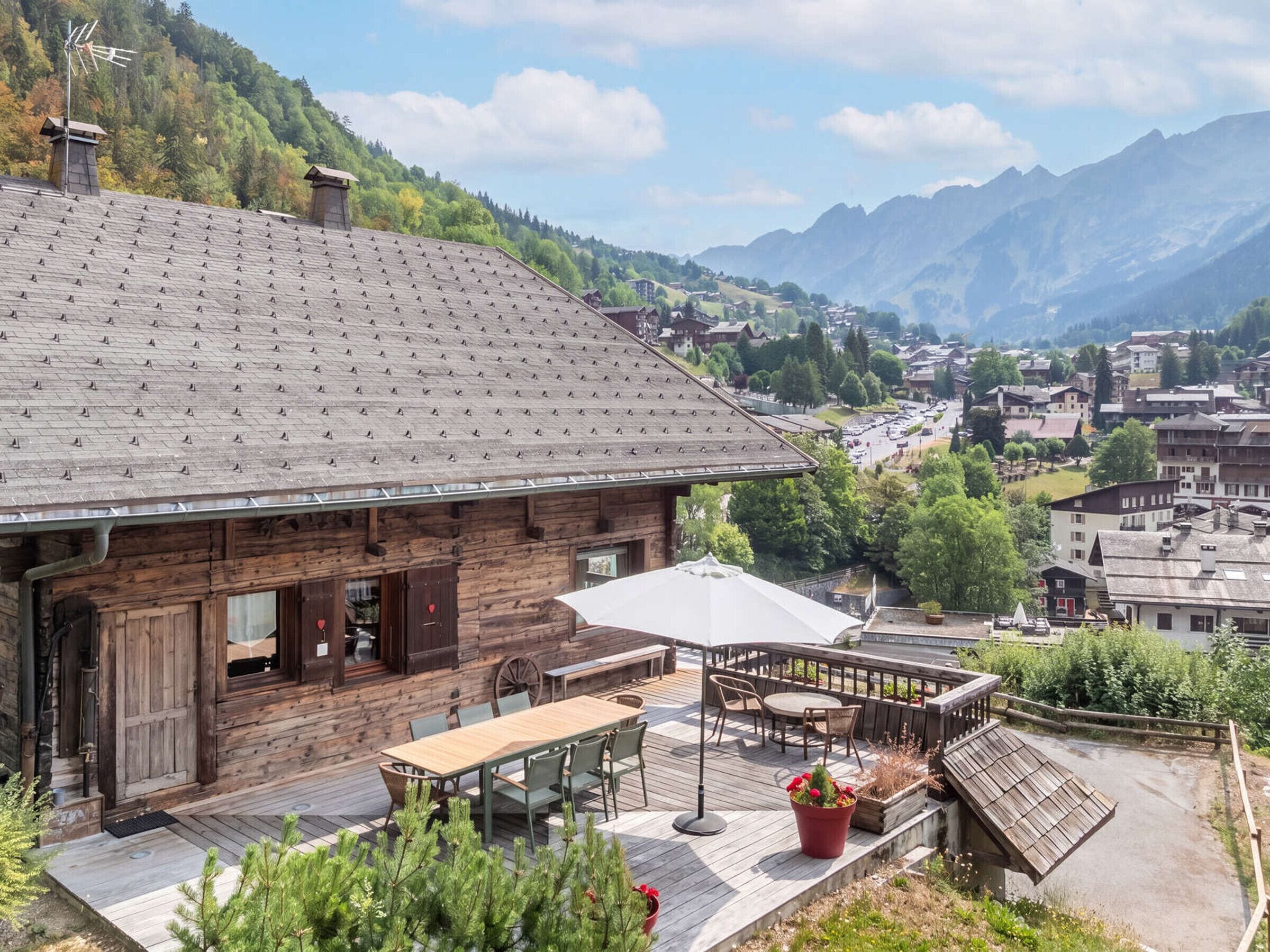 Una cabaña de madera con terraza, mobiliario exterior y vistas a un pueblo de montaña.