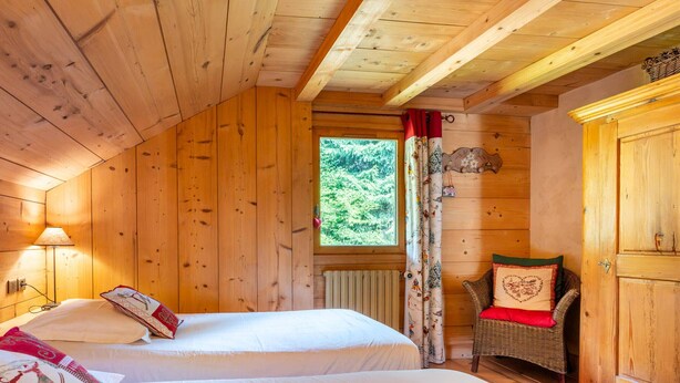 Quarto de cabana de madeira com duas camas, uma cadeira de vime e uma janela com cortinas.