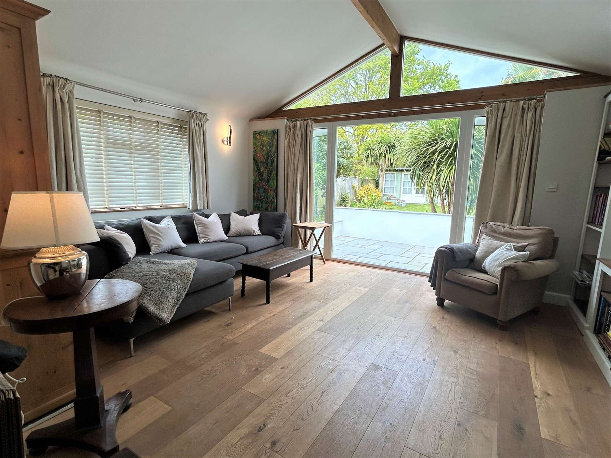 A spacious living room with a large sofa, a smaller armchair, a coffee table, and a lamp. There is a sliding glass door leading to a patio with a view of greenery.