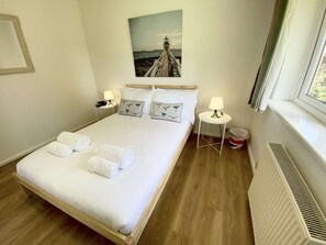 A hotel room with a bed, two bedside tables, a window, a radiator, and a painting of a lighthouse. - Daisy Cottage by NewFo dot Rest (Lymington)