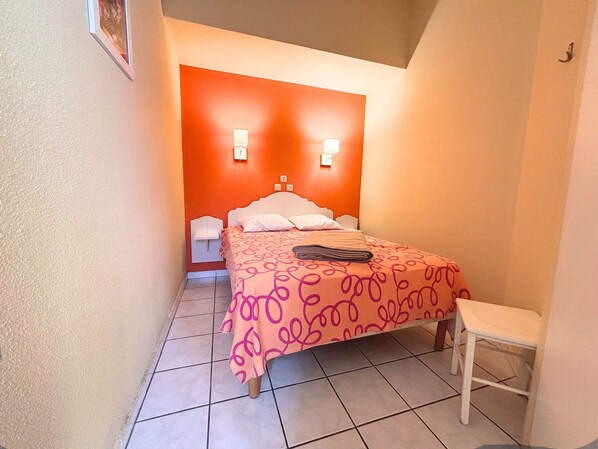 Een kleine, smalle hotelkamer met een bed, een nachtkastje, een stoel en twee wandlampen.