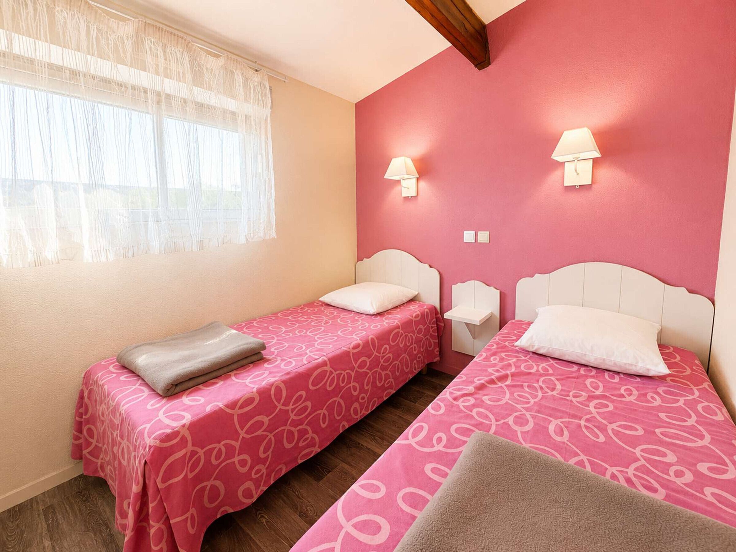 Quarto com duas camas, cada uma com colcha estampada em rosa e cabeceiras brancas. Há duas luminárias de mesa e uma janela com cortinas finas.