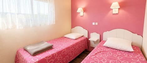 Habitación con dos camas, cada una con una colcha rosa estampada y cabeceros blancos. Hay dos lámparas de noche y una ventana con cortinas traslúcidas.