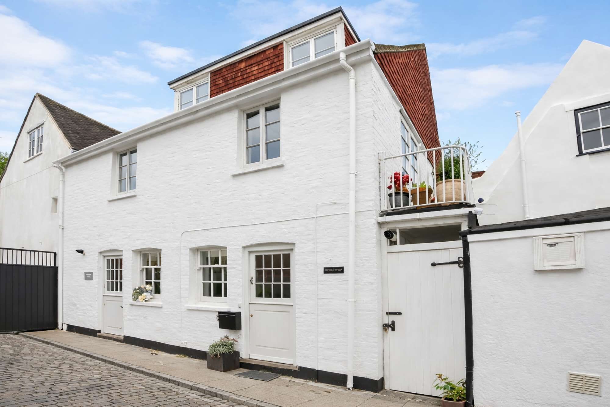 A white building with a black gate, a small balcony with potted plants, and a cobblestone street.