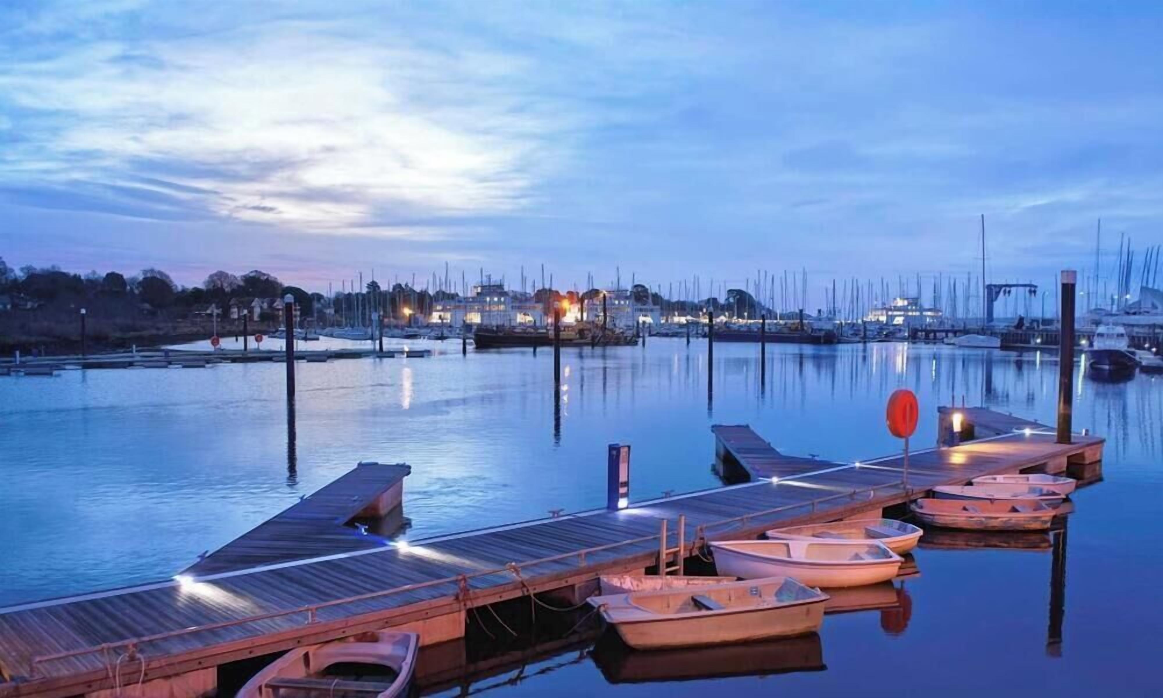 Un port de plaisance avec des bateaux amarrés à des quais en bois, une eau calme et une vue sur la ville en arrière-plan.