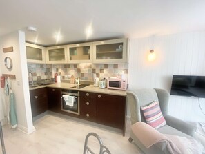 A compact kitchen with a microwave, toaster, and kettle, a built-in oven, and a dining area with a TV and a sofa. - Newgate Cottage by NewFo dot Rest (Lymington)
