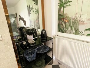 Una màquina de cafè sobre un taulell negre amb un radiador blanc i un mural tropical a la paret.