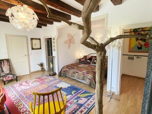 Una habitació amb un llit, una cadira, una catifa colorida, un arbre decoratiu i una làmpada penjant.