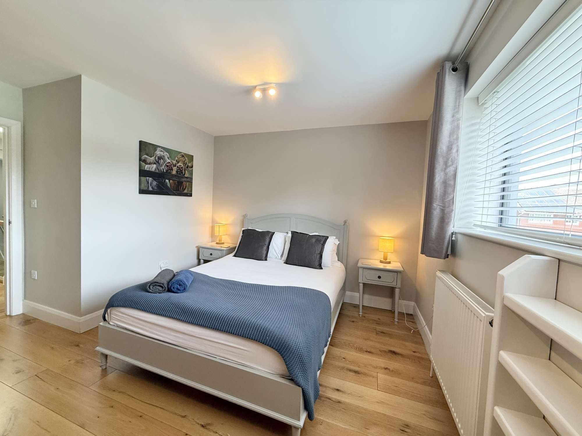 A bedroom with a bed, bedside tables, a window with blinds, and a painting on the wall.