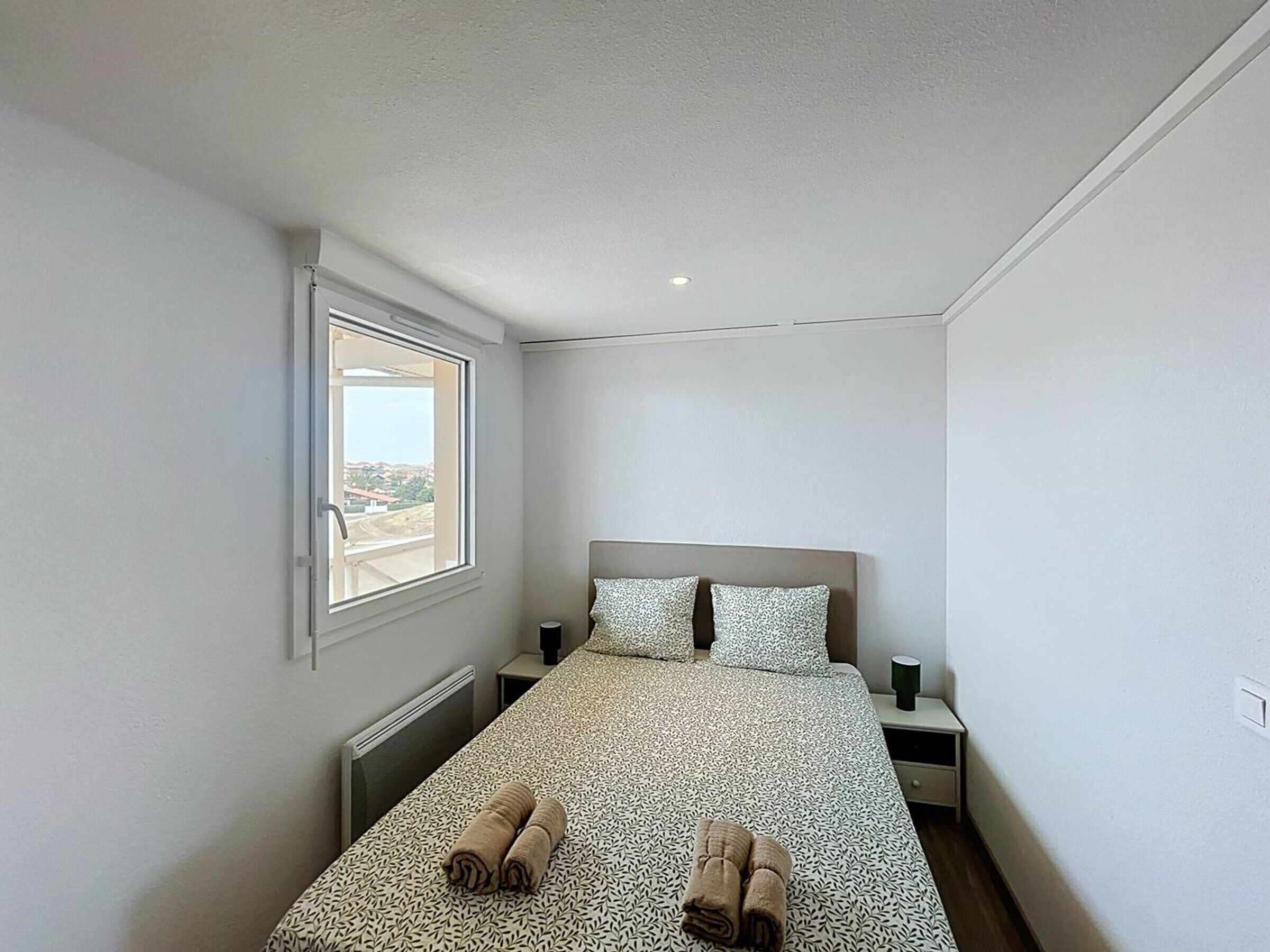 Ein Schlafzimmer mit einem Bett, zwei Kissen, einem Nachttisch, einem Fenster und Blick ins Freie.