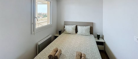 Ein Schlafzimmer mit einem Bett, zwei Kissen, einem Nachttisch, einem Fenster und Blick ins Freie.