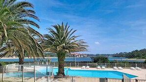 Una piscina con sillas de descanso, palmeras y vistas al mar y edificios al fondo.
