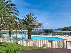 Ein Schwimmbecken mit Liegestühlen, Palmen und Blick auf das Meer sowie Gebäude im Hintergrund.