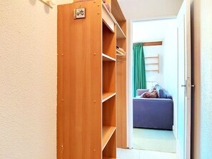 Ein Holzschrank mit Regalen und Kleiderstange, der zu einem Raum mit einer Couch und einem grünen Vorhang führt.