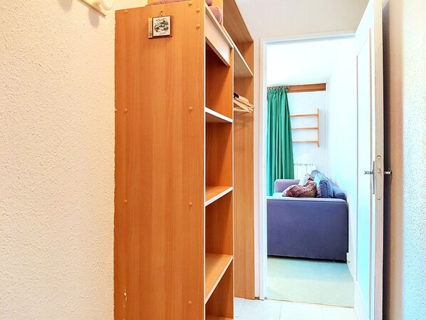 Een houten kast met planken en een kledingroede, die leidt naar een kamer met een bank en een groen gordijn.