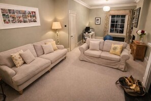 Ein Wohnzimmer mit zwei beigefarbenen Sofas, einer Lampe, einer gerahmten Fotocollage und einem Fenster mit Vorhängen.