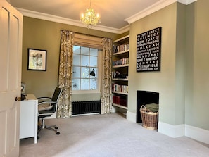 Ein Zimmer mit Heizkörper, Bücherregal, Schreibtisch mit Stuhl, Fenster mit Vorhängen und einer Chandelier.