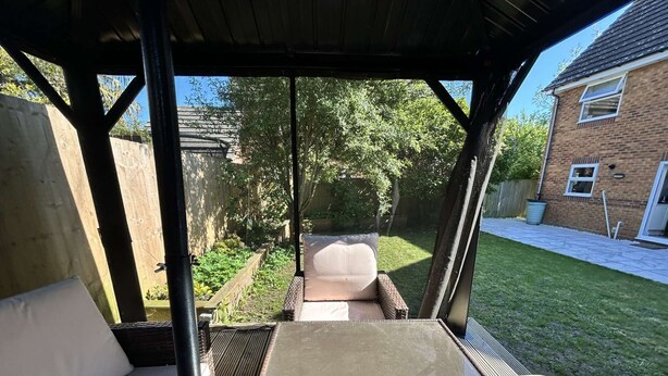 Un patio techado con una mesa y dos sillas, con vista a un jardín que tiene césped y una casa de ladrillo.
