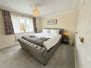 A bedroom with a large bed, a bedside table with a lamp, a radiator, and a window with curtains. - Vitre Gardens by NewFo dot Rest (Lymington)