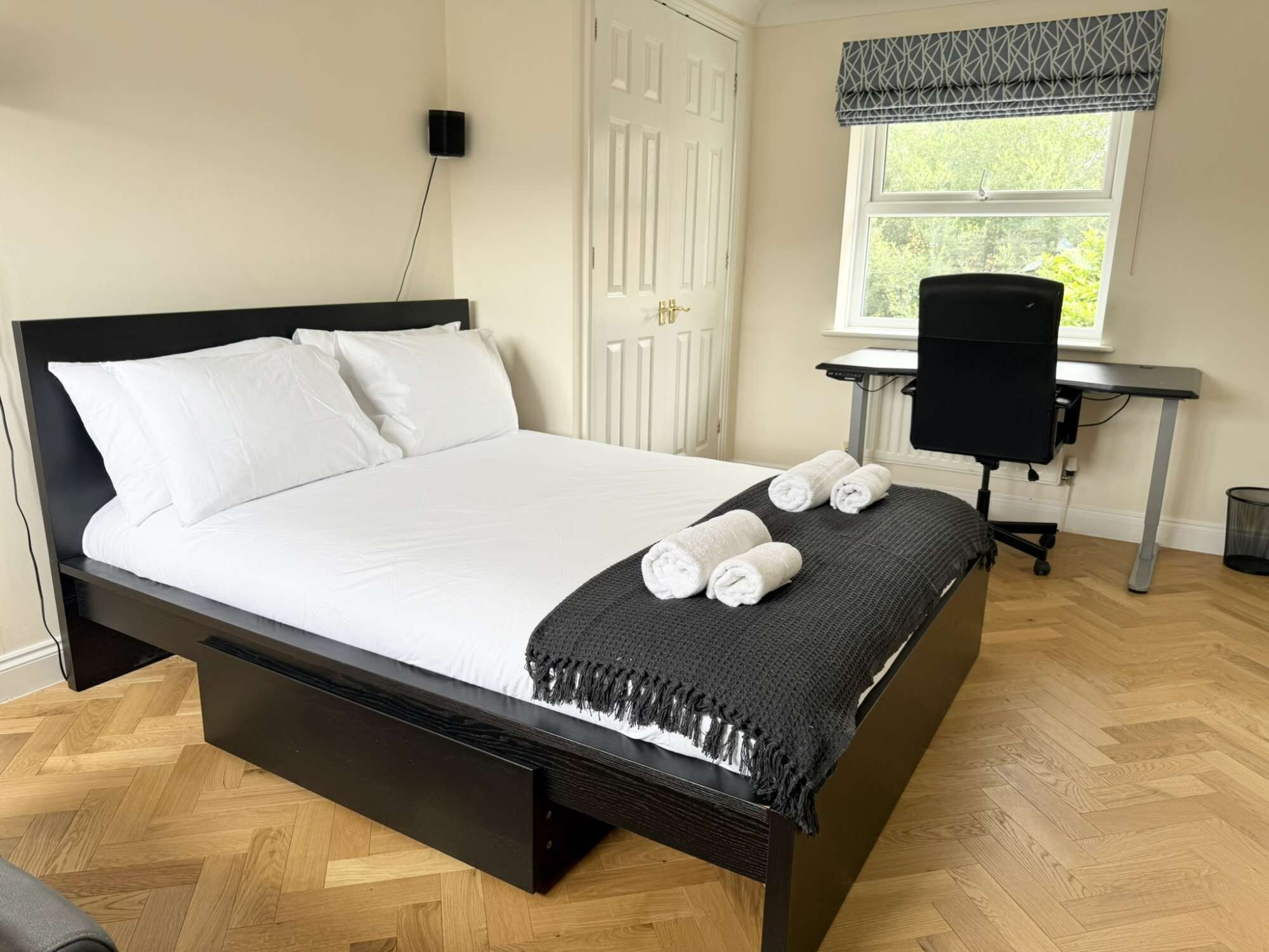 A neatly made bed with white linens and a dark headboard, a small desk with a chair, and a window with a view of greenery.