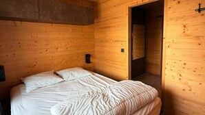 Puisesta hirrestä rakennettu huone, jossa on sänky, pieni pöytä ja ovi, joka johtaa toiseen huoneeseen.