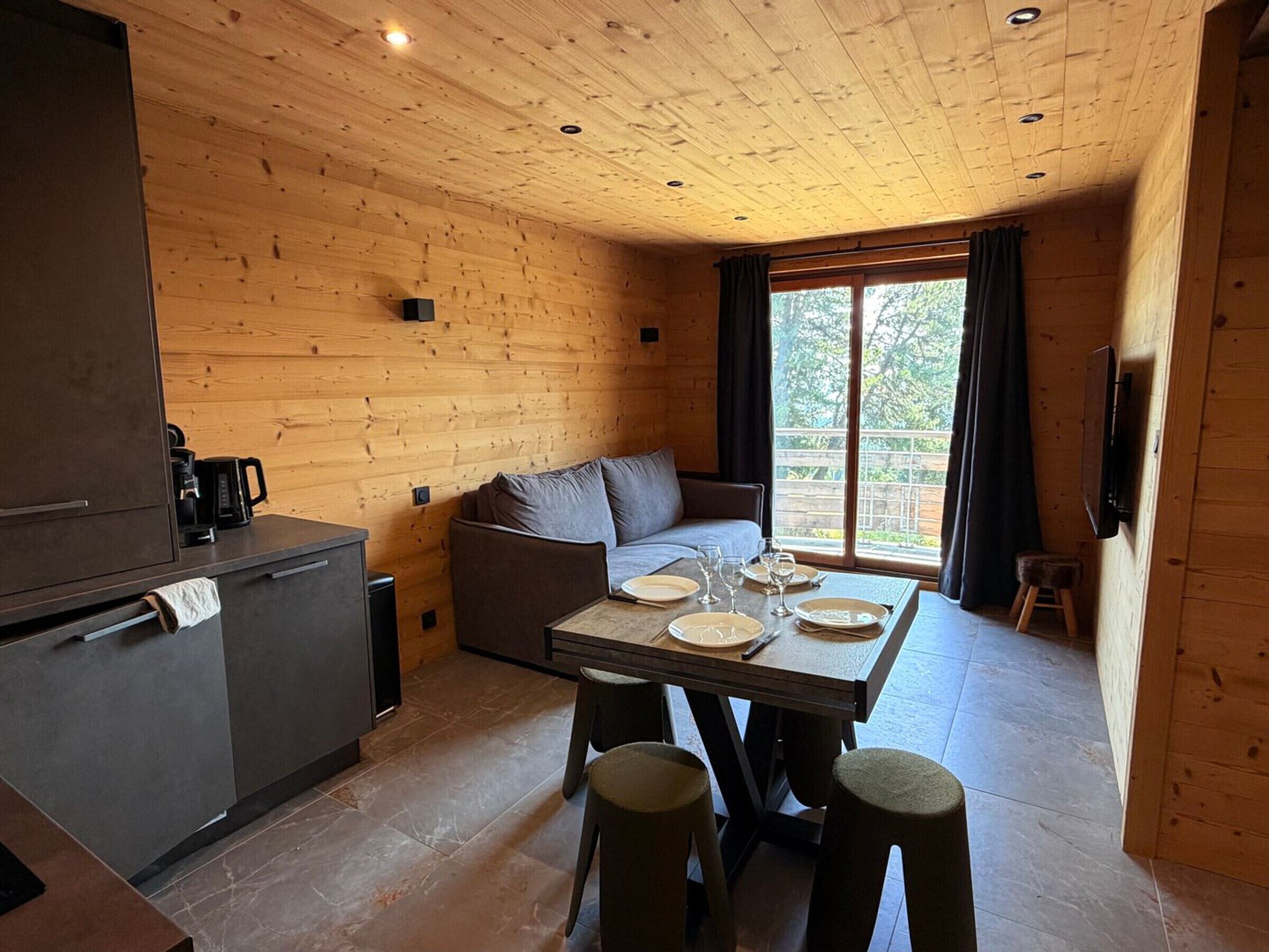 A cozy cabin interior with a wooden interior, a sofa, a dining table set for two, and a kitchen area.