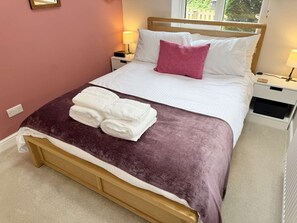 A neatly made bed with a purple blanket, white linens, and a pink pillow. There are two bedside tables with lamps, a power outlet, and a window with a view of greenery. - Yew Tree Cottage – Direct New Forest Access, Ponies & Walk to Lyndhurst (Lyndhurst)