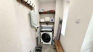 A laundry room with a washing machine, dryer, ironing board, and a bench. - Yew Tree Cottage – Direct New Forest Access, Ponies & Walk to Lyndhurst (Lyndhurst)