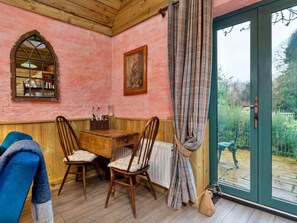 A cozy room with a wooden table and chairs, a radiator, a window with a plaid curtain, and a green door leading outside. - Countryside Barn Near River Great Ouse (Huntingdon)