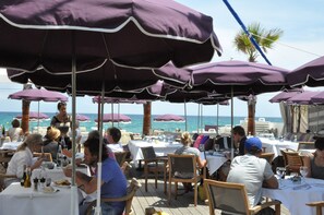Un espace extérieur aménagé pour les repas, avec des tables et des parasols violets, offrant une vue sur la mer.