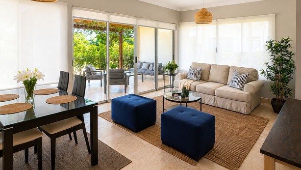 A modern living room with a glass door leading to an outdoor area, a sofa, ottomans, a coffee table, and a dining table with placemats. - Room-luxury Hacienda Residence Punta Can (Punta Cana)
