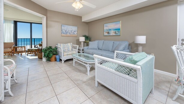 Amplia sala de estar con balcón y vista al mar, amoblada con muebles de mimbre blanco y decorada con dos cuadros.