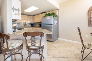 A kitchen with a refrigerator, a coffee maker, and a plant on top. There are two chairs with a decorative backrest. - Phoenix X 908 1 Bedroom Condo (Orange Beach)